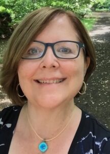 Nicole Hennig with light brown, chin-length straight hair wearing a necklace with bright blue pendant, a dark shirt with white patterns, and square eyeglasses with black frames. Trees and nature walking paths behind her.