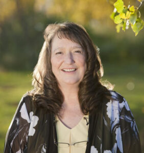 Woman with shoulder lenght wavy brown hair wearing light yellow short with black and white overtop. She's smiling at the camera.