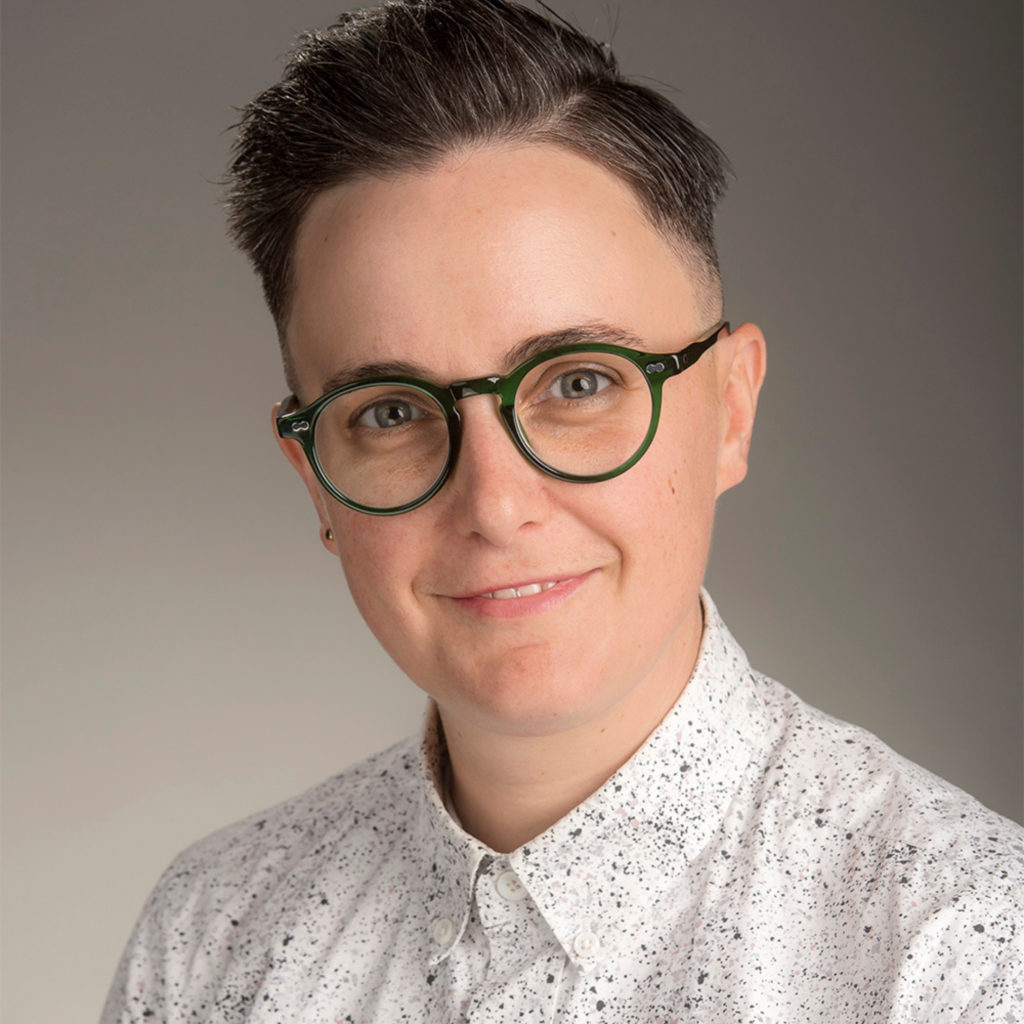 A person with short hair and green glasses smiles for a portrait