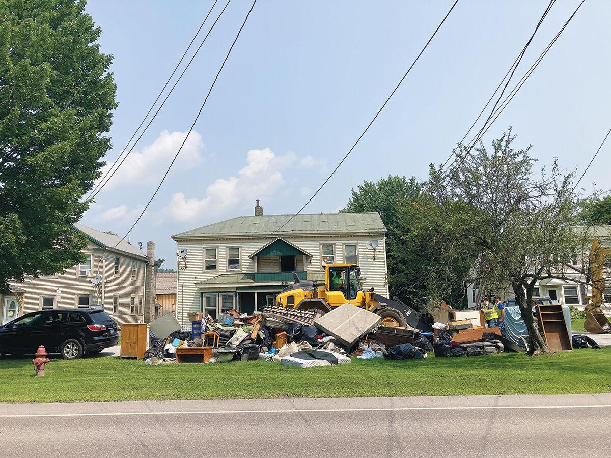 Color photo of across the street view of three homes with flood soaked content and debris in large pile on yard.