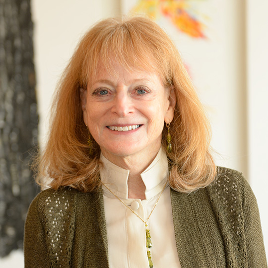 Color headshot photo of Carol Mandel wearing olive green sweater, ivory shirt, standing near tree in front of ivory building.