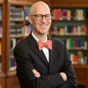 Color image of John Wilkin wearing dark suit with orange bow tie, standing in front of library shelves