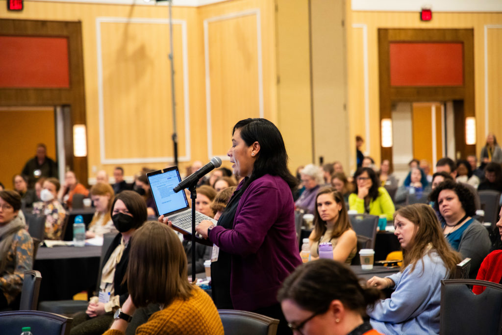 Person at microphone in center of room with folks seated at tables, holding a laptop