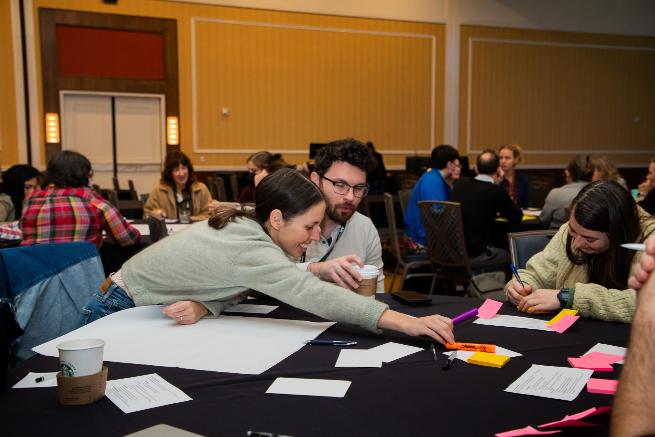 People seated at a roundtable with papers and markers. One person reaching for a marker.